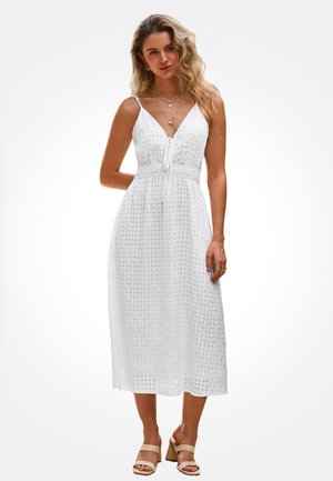 Kobieta ubrana w białą, bezrękawnikową sukienkę midi z teksturowanym wzorem w kratę oraz beżowe sandały na obcasie, ozdobiona warstwowymi naszyjnikami i pierścionkami.