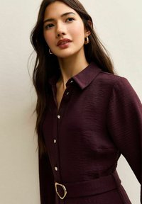 Camicia con bottoni color amaranto con finitura testurizzata, dotata di colletto, leggero effetto lucido e vita con cintura e fibbia metallica rotonda.