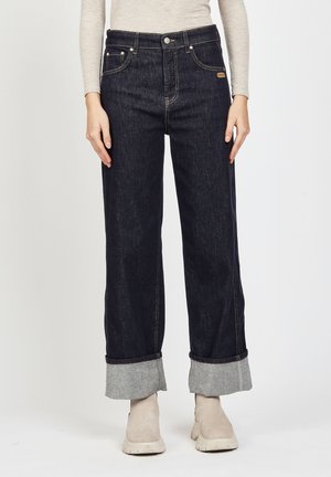 Person wearing dark blue cuffed wide-leg jeans with contrast stitching, beige top, and light beige ankle boots standing against a plain background.
