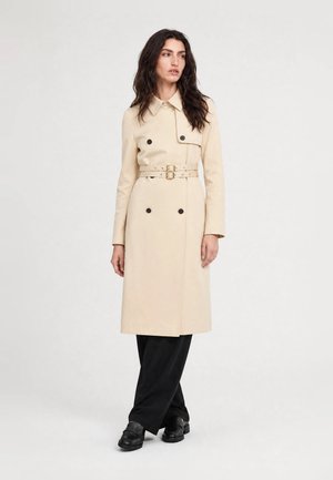 Mujer con gabardina beige con cinturón, pantalones negros de pierna ancha y mocasines negros, de pie frente a un fondo claro y liso.