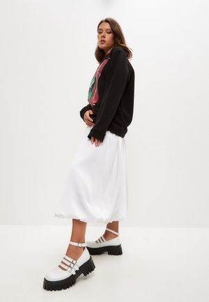 Felpa nera con stampa grafica, gonna midi plissettata bianca e scarpe con piattaforma bianche con più cinturini e suole nere spesse.