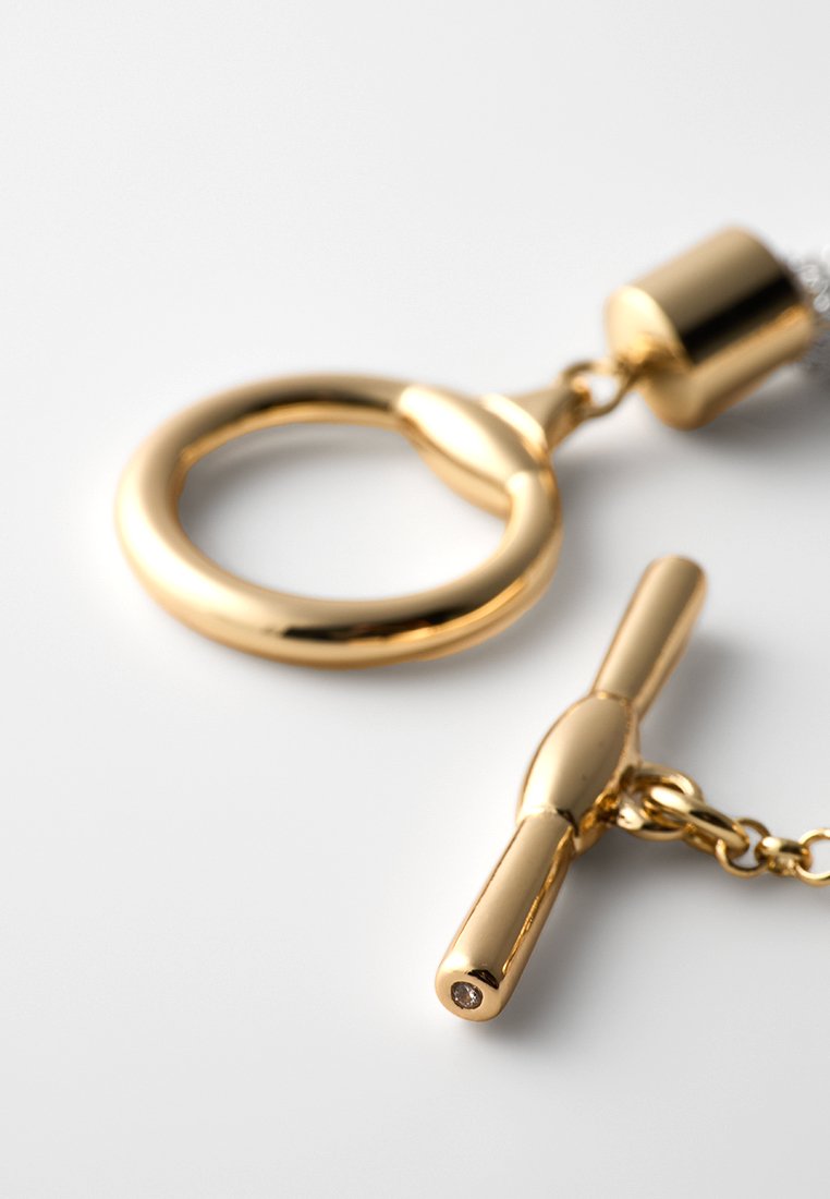 Gros plan sur un fermoir à bascule en or brillant sur un collier, présentant un anneau circulaire et une barre horizontale avec une petite pierre précieuse intégrée.