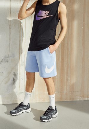 Persoon die een zwart Nike tanktop draagt, lichtblauwe Nike shorts, witte Nike sokken en zwarte Nike sneakers, staand tegen een textuurmuur.