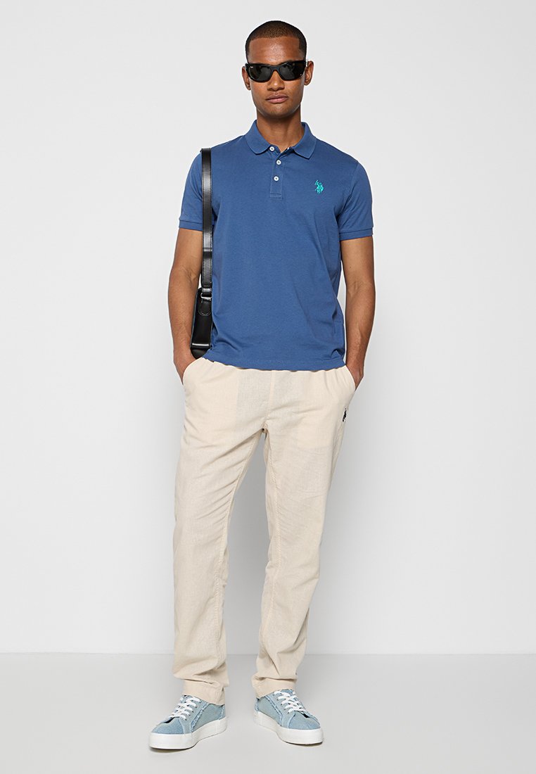 Blaues Poloshirt mit grünem Logo, beigefarbene Leinenhose und hellblaue Sneakers. Modell trägt schwarze Sonnenbrille und steht mit einer schwarzen Tasche.