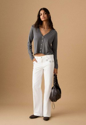 Mujer con un cárdigan gris corto abotonado, pantalones blancos con detalles de estrellas, sosteniendo un bolso negro fruncido sobre un fondo beige.