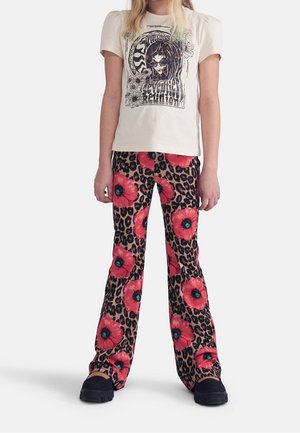 Enfant portant un t-shirt graphique beige et un pantalon imprimé léopard avec de grandes fleurs rouges, debout avec les bras détendus sur les côtés.