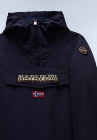 Tengerészkék pulóverdzseki magas nyakkal, félig cipzáras záródással, elölvarrt zsebbel és hímzett zászlókkal; márkázott logó díszítésekkel.