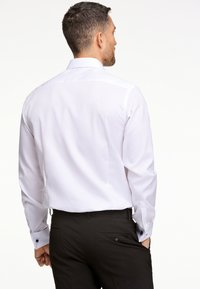 Camicia bianca con maniche lunghe, polsini con bottone e colletto classico, abbinata a pantaloni neri. Tessuto morbido con vestibilità su misura.