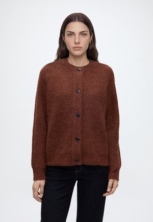 Femme aux cheveux bruns et lisses porte un cardigan tricoté marron boutonné et un pantalon foncé, debout devant un fond clair uni.