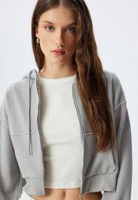 Sudadera gris con cremallera y textura acanalada, dos bolsillos frontales y capucha con cordón, llevada sobre una camiseta crop blanca lisa.