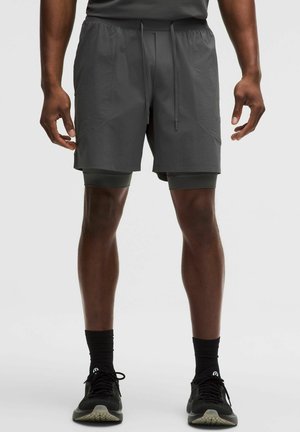 Homme portant un short de sport à double couche gris foncé, des chaussettes noires et des chaussures de course noires, debout devant un fond blanc.
