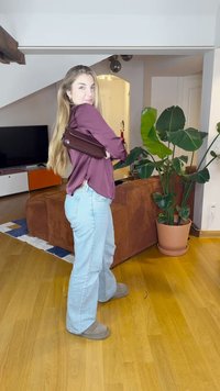 Bordó blůza, světle modré zvonové džíny a šedé pantofle. Tmavě hnědá taška je přehozená přes jedno rameno. Vedle stojí zelená rostlina.