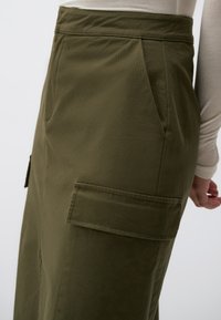 Olijfgroene rok met een hoge taille, voorzien van plooien en twee grote, platte zakken. Gemaakt van een lichtgewicht stof met een gladde textuur.
