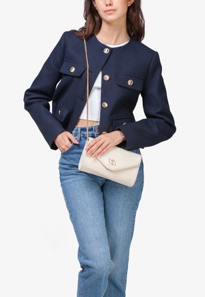Femme portant une veste bleu marine à boutons, un crop top blanc, un jean bleu, tenant un sac à main blanc avec une chaîne dorée portée en travers du corps.