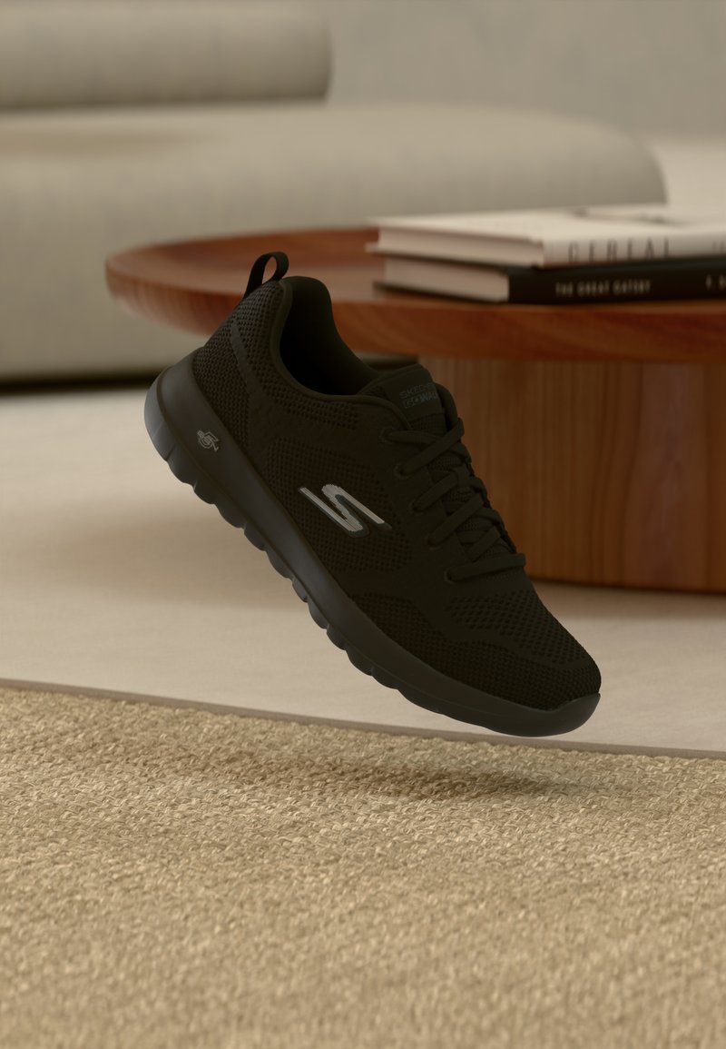 Zapatilla deportiva negra con parte superior de malla, patrón texturizado, cierre de cordones y suela acolchada. Cuenta con un logotipo en el lateral.