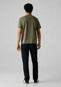 Olivno zelena majica s kratkimi rokavi in okroglim izrezom, z logotipom na hrbtu, v kombinaciji s temnimi jeans hlačami in sproščenimi čevlji.