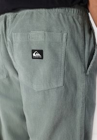 Pantaloni din corduroy de culoare verde deschis, cu o suprafață texturată, prezentând buzunare laterale și un buzunar la spate cu o etichetă patch Quiksilver de culoare neagră.