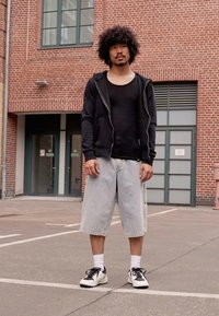 Hanorac cu fermoar negru, maiou negru aderent, pantaloni scurți blegi ușor, șosete albe, adidași negri și albi, stând pe trotuar lângă un zid de cărămidă.