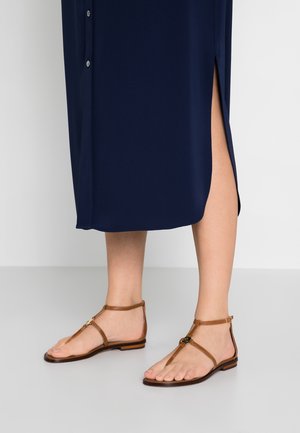 Brązowe płaskie sandały z zapięciem T. Posiadają cienki pasek wokół kostki oraz małe metalowe akcenty. Podeszwa jest płaska i lekko teksturowana.