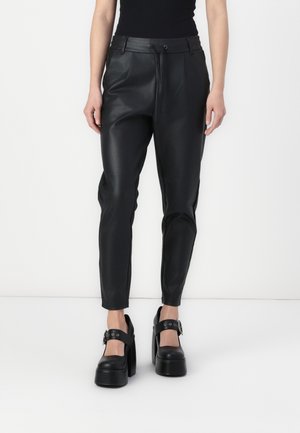 Persoon die zwarte hooggetailleerde leren broek met taps toelopende pijpen draagt en zwarte platform Mary Jane schoenen met zilveren gespsluitingen.