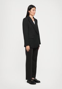 Svart dobbeltradet blazer med notched lapels, supplert med rette svarte bukser og blanke svarte loafers. Glatt stofftekstur.