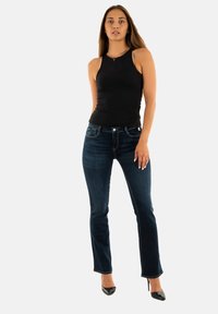 Top negro sin mangas, jeans rectos azul marino y zapatos de tacón altos negros. La modelo está de pie en posición recta frente a un fondo blanco.