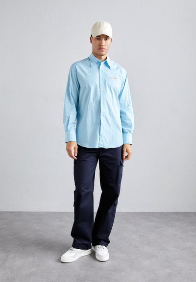 Camisa de botón a rayas azules, pantalones cargo azul marino y zapatillas blancas. Presenta mangas largas, un cuello y un detalle del logo en la parte frontal.