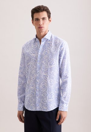 Mężczyzna ubrany w jasnoniebieską, kwiecistą koszulę na guziki z długim rękawem oraz ciemne spodnie, stojący na tle jednolitego tła.