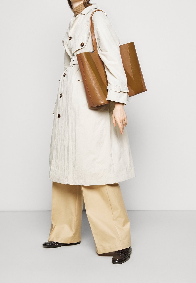 Personne portant un trench-coat beige et un pantalon à jambes larges tenant un grand sac cabas en cuir marron sur l'épaule, portant des mocassins foncés.
