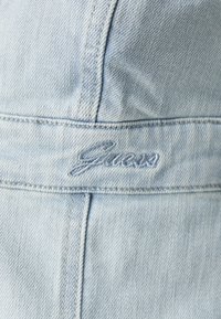 Lyseblå denimjakke med teksturert overflate, brodert "Guess"-logo og kontrasterende sømmer langs stingene.