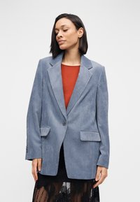 Blauwe corduroy blazer met een kraag met inkeping, enkele knoopsluiting, twee voorzakken en een gestructureerde stof. Gedragen over een roestkleurige top.