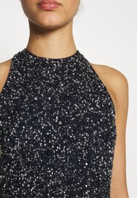 Navy sininen ilmanhiha dressi, jossa on korkea kaulus, koristeltu tiheällä kerroksella hopea- ja harmaasävyisiä paljetteja, jotka luovat teksturoidun ja hohtavan vaikutuksen.