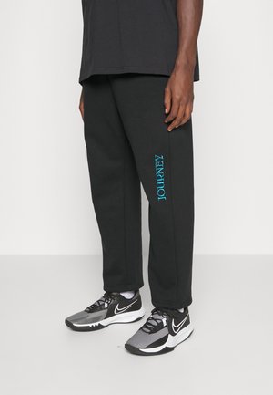 Zwarte joggingsbroek met een verticale "JOURNEY" graphic in het blauw op het linkerpoot, gecombineerd met grijze en zwarte sportschoenen.