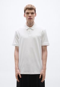Fehér, sima pamutból készült galléros póló, klasszikus gallérral, két gombbal és finom márkajelzéssel a bal mellrészen. Egyszerű kialakítás.