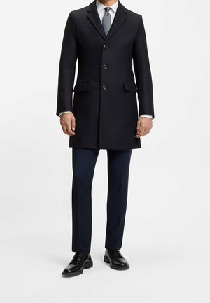 Homme portant un pardessus noir boutonné, une chemise blanche, une cravate bleue, un pantalon foncé et des chaussures habillées noires, debout sur fond blanc.