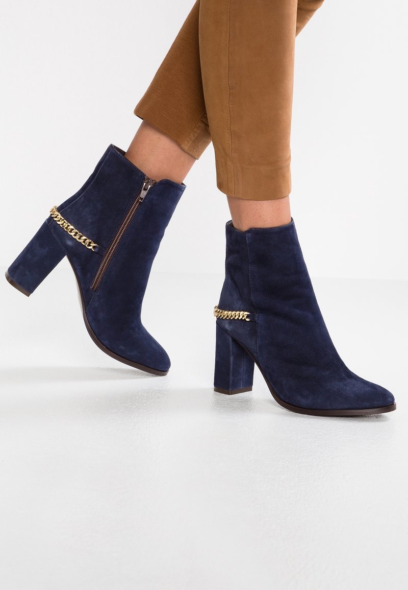 Bottines en daim bleu marine avec un bout carré, un talon épais et un détail de chaîne dorée autour de la cheville. Fermeture zippée sur le côté intérieur.