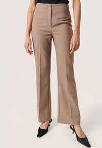 Beige mukautetut housut suoran lahkeen muotoilulla, etuvetoketjulla ja kulutustiedoilla, yhdistettynä mustiin slingback-korkokengiin.