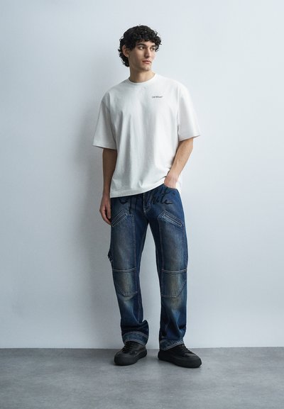 Νέος άνδρας που στέκεται, φορώντας λευκό T-shirt Off-White, μπλε τζιν με μπαλώματα και μαύρα γράμματα, και μαύρα αθλητικά παπούτσια, μπροστά σε απλό γκρι τοίχο.