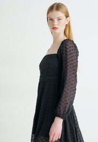 Czarna koronkowa sukienka z dopasowanym gorsetem i długimi, przezroczystymi bufkami. Tkanina ozdobiona jest wzorami kwiatowymi i falistym pasem w talii.