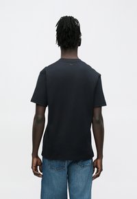 Άνδρας με κράνμπακς που φοράει μαύρο κοντομάνικο πουκάμισο και μπλε τζιν, στέκεται πλάτη στην κάμερα μπροστά σε απλό φόντο.