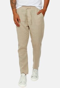 Pantaloni beige di lino con elastico in vita e cordoncino, caratterizzati da una vestibilità rilassata e gambe affusolate, abbinati a sneaker bianche.