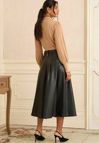 Barna kötött felső puffos ujjakkal, fekete, pliszírozott műbőr midi szoknyával párosítva. Fekete magassarkú cipők egészítik ki az öltözetet.