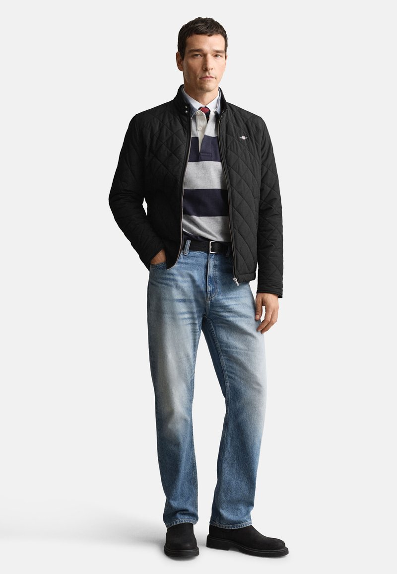 Sort quiltet jakke, grå og marineblå stribet polo, lyseblå jeans og sorte støvler. Enkel design med lynlåslukning og uden synlige logoer.