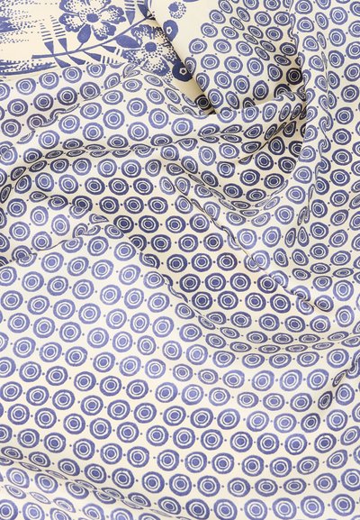 Λευκό ύφασμα με μπλε κυκλικά και αφηρημένα λουλουδένια σχέδια, απαλά διπλωμένο ώστε να αναδεικνύει την υφή και τις λεπτομέρειες του σχεδίου.
