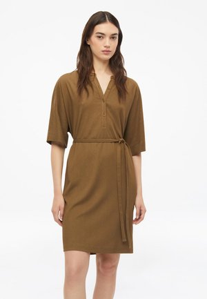 Femme portant une robe marron mi-longue avec des manches mi-longues, un col en V boutonné et une ceinture nouée à la taille sur fond blanc.
