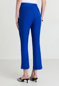 Pantaloni blu con una texture liscia, vita media e design a gamba dritta. Presentano fessure laterali all'orlo. Indossati con mules con tacco bianchi e neri.