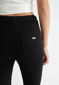 Melnas augsto jostu džinsus ar gludu tekstūru, divām aizmugures kabatām un nelielu metalizētu logotipa akcentu labajā kabatā.