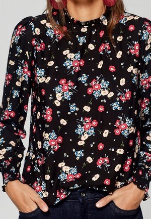 Femme portant un chemisier noir à manches longues avec des motifs floraux rouges, bleus et blancs, associé à un jean bleu foncé et des boucles d’oreilles à pompons roses.