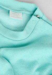 Mintunvihreä vauvan neulepaita, jossa on resorikaulus, nepparikiinnitys ja pehmeä kangas. Labelissa lukee "NEXT baby", valmistettu Intiassa.