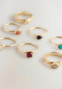 Achteteiliges Set aus goldenen Ringen mit verschiedenen Steinen: rot, schwarz, türkis und weiß. Unterschiedliche Formen, darunter oval, rund und quadratisch. Strukturierte Akzente.